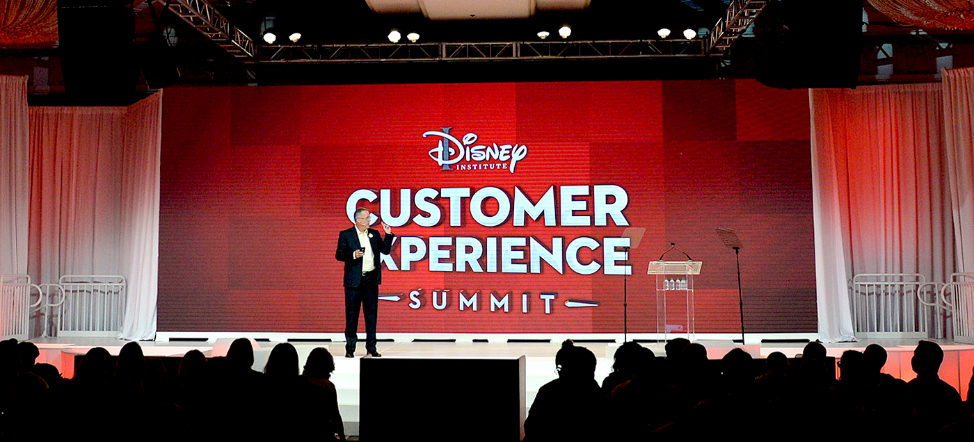 man standing on stage with background  text Disney Institute Customer Experience Summit